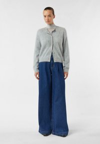 Jasnoszary kardigan z wełny z zapięciem na guziki z przodu, ściągaczami na rękawach, zestawiony z szerokimi, niebieskimi dżinsami z wysokim stanem. Prosty, teksturowany design.