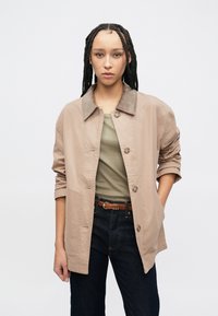 Šviesiai ruda mygtukais užsegama striukė su korduro dildu, pasižyminti laisvu kirpimu ir suvertomis rankovėmis, derinama su aptemptu alyvuogių spalvos viršutiniu drabužiu ir tamsiomis džinsinėmis kelnėmis.