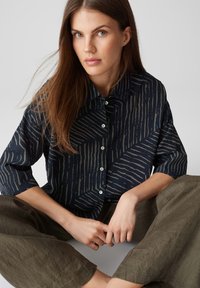 Navyblaues Hemd mit kontrastierendem beige gestreiften Muster, geknöpfter Front und kurzen Ärmeln, kombiniert mit lockeren olivgrünen Hosen.