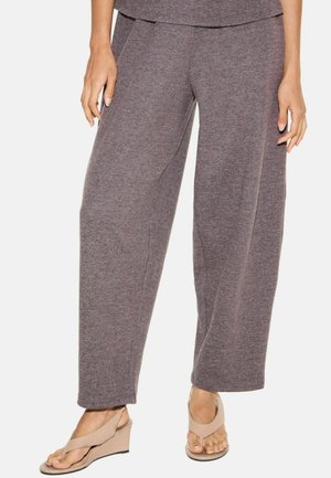 Femme portant un pantalon gris ample et des sandales compensées beiges, debout devant un fond uni.