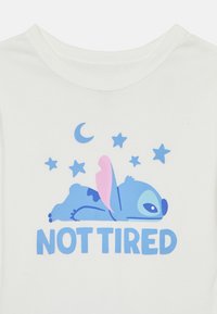 T-shirt en coton blanc avec un personnage de dessin animé bleu allongé, agrémenté d'accents roses, entouré d'étoiles et d'une lune croissante, avec le texte « PAS FATIGUÉ. »