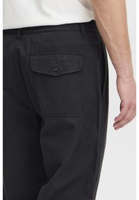Black cotton trousers featuring a back pocket with a button closure and an elastic waistband for comfort. Visible stitching detailing.