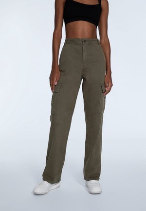 Personne portant un pantalon cargo vert olive, des baskets blanches et un débardeur court noir, debout devant un fond uni.