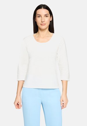 Biały, prążkowany top z dekoltem w kształcie litery U i rękawami do połowy przedramienia, noszony z jasnoniebieskimi spodniami. Gładka faktura z subtelnymi poziomymi liniami.