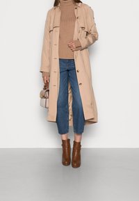 Beżowy trencz z guzikami, brązowy sweter z prążkami, niebieskie krótkie dżinsy i brązowe botki za kostkę. Mniejsza torba trzymana w ręku.