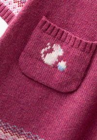 Megzta megėlė turtingu rožiniu atspalviu, su kišeniu, kuriame pavaizduota balta katė ir mėlyna kiaušinio dekoracija. Minkšta tekstūra su vagoto dizaino detalėmis.