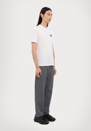 Camiseta de algodón blanca con un parche de logo negro, combinada con unos pantalones vaqueros de denim gris de corte holgado y zapatillas negras, mostrada sobre un fondo neutro.