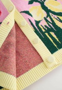 Villainen peitto, jossa on kukkakuvio pinkin, keltaisen ja vihreän sävyissä, teksturoitu keltainen ribbineulosreuna ja napitettu yksityiskohta yhdellä sivulla.