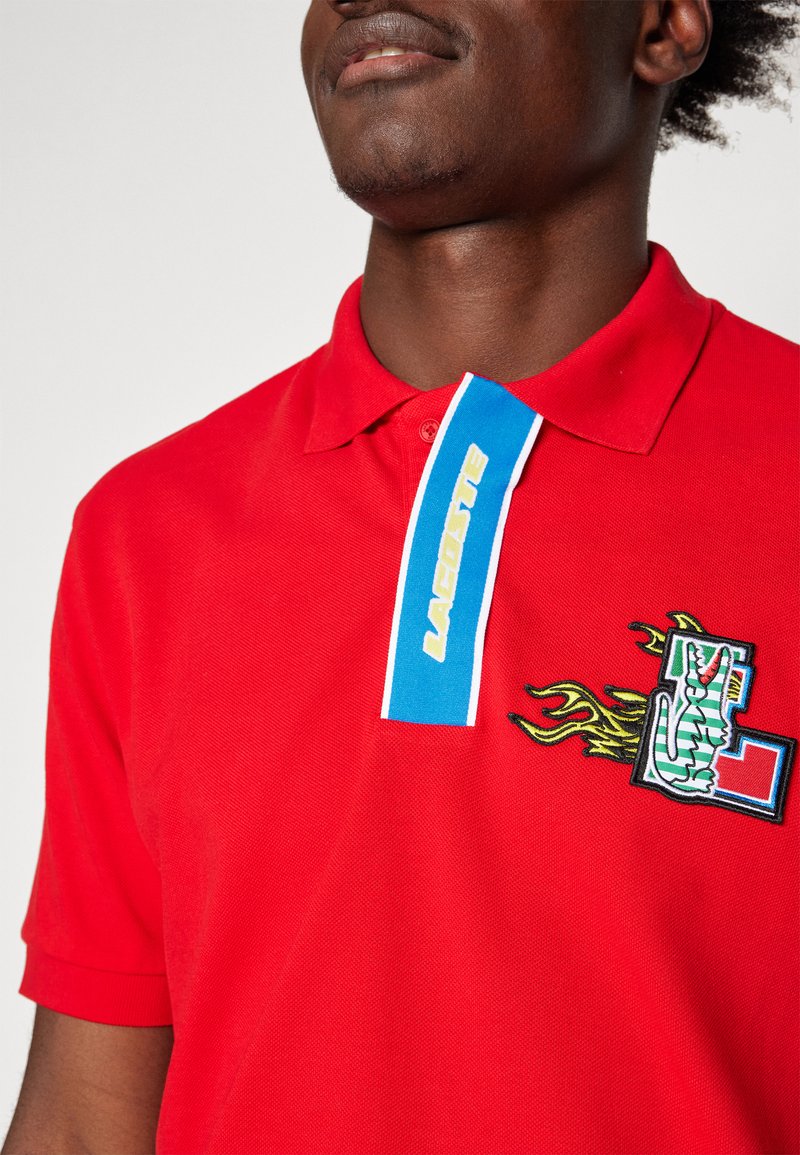 Polo rouge avec un col bleu et blanc, arborant un logo brodé et un design de flamme. Tissu texturé avec une coupe classique.