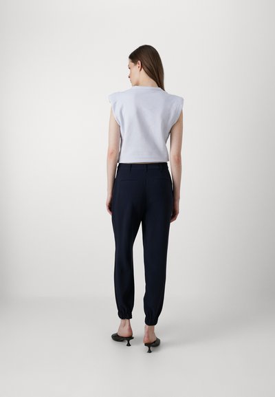 Witte kortekleurige top met schouderkussens, gecombineerd met marineblauwe tapered broek met elastische manchetten en zijzakken. Zwarte hakken mules.