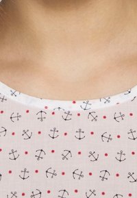 Biały materiał z powtarzającym się wzorem niebieskich kotwic i czerwonych kropek; okrągły dekolt i gładka tekstura.