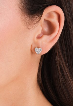 Oreille d'une femme portant une petite boucle d'oreille en argent en forme de cœur, incrustée de cristaux transparents, sur des cheveux brun foncé.