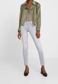 Veste en suédine olive avec des accents texturés sur les épaules, associée à un jean skinny bleu clair et des escarpins noirs à bout pointu.
