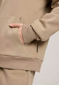 Beżowa bluza z kapturem, wykonana z miękkiego materiału, z kangurzą kieszenią, prążkowanymi mankietami oraz małym czarnym akcentem przy szwie bocznym.