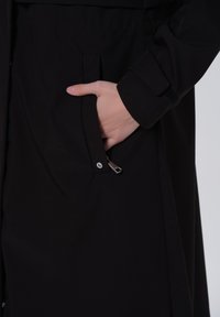 Cappotto nero con una texture liscia, dotato di una tasca laterale con chiusura a zip e dettagli di polsini in velcro. Design minimalista, senza motivi visibili.