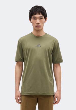 Jeune homme debout, face à l’avant, portant un T-shirt vert olive à manches courtes avec un petit logo sombre sur la poitrine et un pantalon marron.