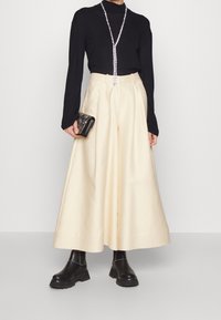 Maglia nera a maniche lunghe, pantaloni crema a gamba larga, collana lucida, pochette nera con decorazioni e stivaletti neri chunky.