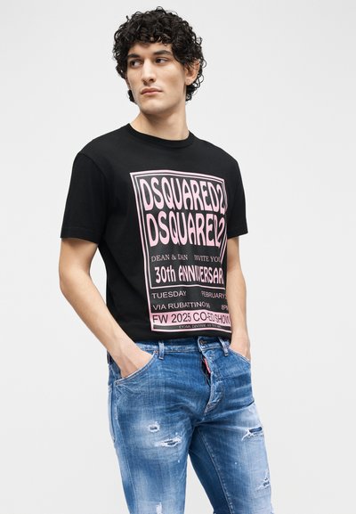 Νέος άνδρας με σγουρά μαλλιά που φοράει μαύρο κοντομάνικο με ροζ γράμματα και σκισμένα μπλε τζιν, με τα χέρια στις τσέπες, κοιτάζοντας προς τα αριστερά.