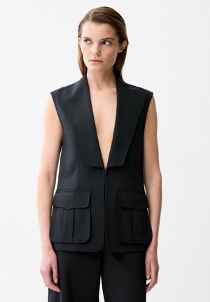 Vrouw met steil bruin haar draagt een mouwloos zwart vest met diepe V-hals en grote klepzakken, kijkt naar links tegen een witte achtergrond.