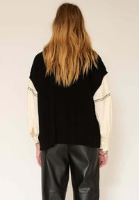 Pull noir surdimensionné à manches courtes superposé sur une blouse crème avec des poignets à motifs, accompagné de pantalons en cuir noir ajustés.
