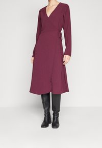 Kvinna i en långärmad, djup V-ringad vinröd omslagsklänning, kombinerad med svarta knähöga klackstövlar, stående mot en enfärgad bakgrund.