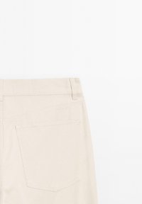 Béžové bavlněné kalhoty s hladkým povrchem, mají zadní kapsu a jemné prošívání podél pasu a okrajů kapes.