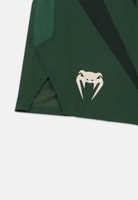 Πράσινο αθλητικό σορτς με υφή υφάσματος, πλαινά πάνελ από δίχτυ και λευκό λογότυπο κόμπρας στο τελείωμα. Διαθέτει πλαϊνή σχισμή για μεγαλύτερη ελευθερία κινήσεων.