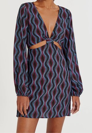 Femme portant une robe courte à manches longues avec des rayures ondulées bleues et violettes, comprenant un profond décolleté en V et un détail découpé à l'avant avec un nœud.
