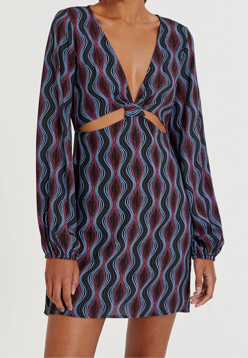Femme portant une robe courte à manches longues avec des rayures ondulées bleues et violettes, comprenant un profond décolleté en V et un détail découpé à l'avant avec un nœud.