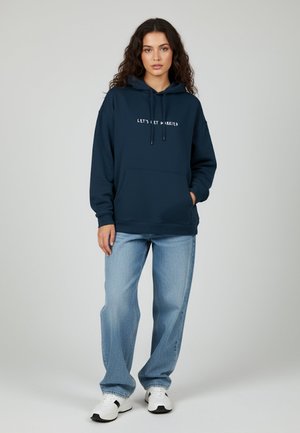 Felpa blu navy con cordoncini regolabili, tasca frontale a marsupio e testo bianco che dice "NUNCIAMO". Abbinata a jeans azzurri e sneakers.