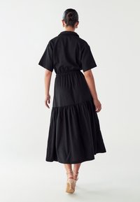 Juoda medvilninė suknelė trumpomis Rankovėmis, su susirinkusia juosmens dalimi ir raukiniais apatiniame krašte. Modelis vilki neutralius sandalus ant aukšto kulno, matoma iš nugaros.