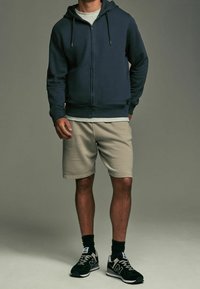 Sweat à capuche zippé marine avec cordons, t-shirt gris clair, short beige et baskets noires avec détails blancs et semelle texturée.