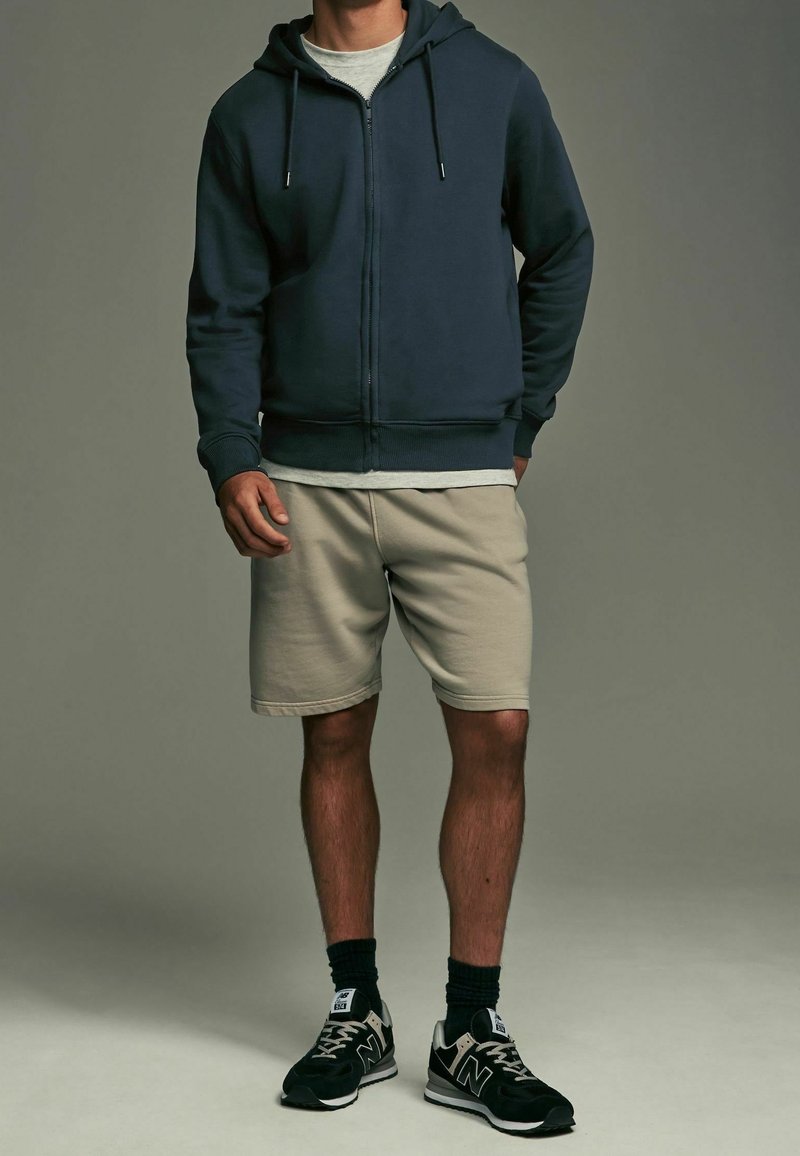Sweat à capuche zippé marine avec cordons, t-shirt gris clair, short beige et baskets noires avec détails blancs et semelle texturée.