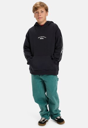 Schwarzer Hoodie mit Logo auf der Vorderseite, langen Ärmeln mit weißen Grafiken, kombiniert mit grünen lässigen Jeans und schwarzen Turnschuhen.