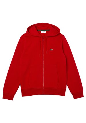 Homme portant un sweat à capuche rouge zippé et un short assorti avec des baskets blanches et rouges, chemise beige en dessous, debout les mains dans les poches.
