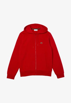 Homme portant un sweat à capuche rouge zippé et un short assorti avec des baskets blanches et rouges, chemise beige en dessous, debout les mains dans les poches.