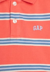 Korallfarget polo skjorte med hvite og blå striper, med krage med to knapper og brodert "GAP"-logo i lys blå nær nederkanten.