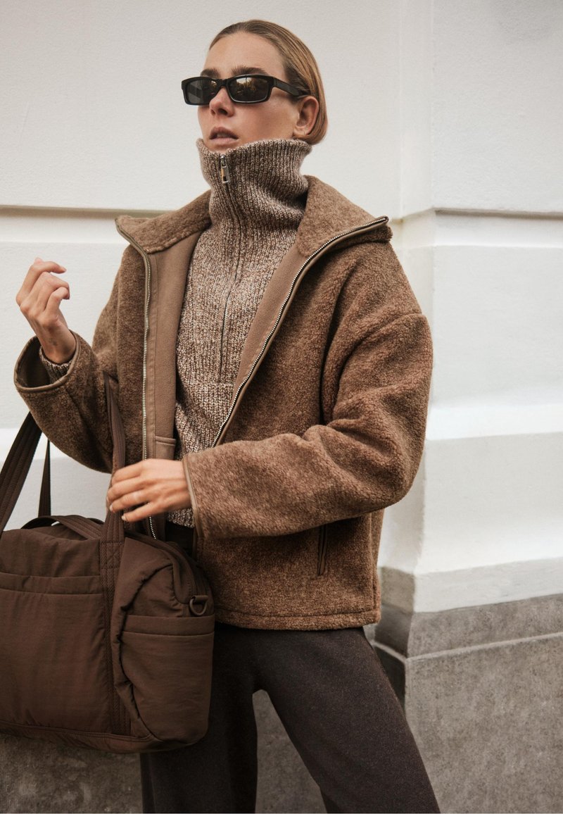 Hnědá fleecová bunda se zipovým límcem, oblečená přes pletený svetr. Černé sluneční brýle a hnědá taška dotvářejí outfit. Viditelné tmavé kalhoty.