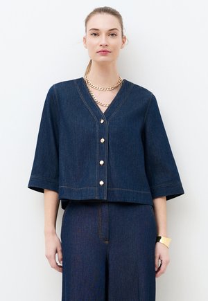 Kobieta ubrana w ciemnoniebieską bluzkę z dżinsu z złotymi guzikami i pasujące szerokie spodnie, uzupełnione warstwowymi złotymi łańcuszkami i złotą bransoletką.