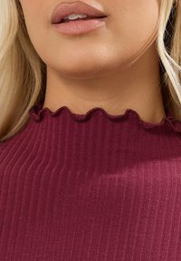 Top bordeaux a coste con colletto alto caratterizzato da un bordo arricciato; texture morbida con strisce verticali e dettagli di design minimalisti.