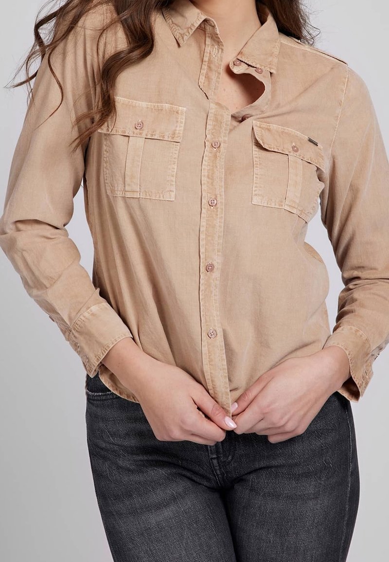 Femme boutonnant une chemise à manches longues marron clair avec poches poitrine, associée à un jean gris foncé, sur un fond gris clair.
