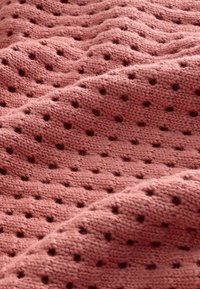 Tekstūruotas megztas audinys švelnios rožinės spalvos su mažu skylučių raštu, turintis bangų formą ir pūkuotą, jaukų išvaizdą.