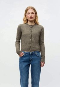 Cardigan castanho ajustado, feito de material suave, com decote redondo, fecho de botão e punhos canelados. Combinado com jeans azuis.
