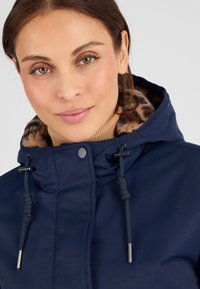 Veste bleu marine avec une capuche doublée en fourrure, dotée de cordons de serrage et de fermetures à pression. Texture lisse avec un léger éclat.