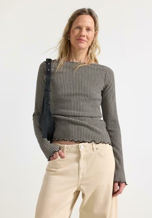 Nainen, jolla on yllään istuva ruudullinen pitkähihainen paita ja beige korkeavyötäröiset housut, kantaa mustaa olkalaukkua seisten yksinkertaista valkoista taustaa vasten.