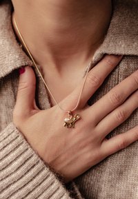 Colgante de oro con forma de elefante en una cadena delgada, que presenta un acento de strass rosa, reposando sobre un suéter texturizado de color marrón claro.