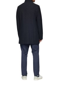 Donker gestreepte wollen jas met een getailleerde pasvorm, hoge kraag en rechte snit, gecombineerd met marineblauwe broek en lichtgekleurde sneakers.