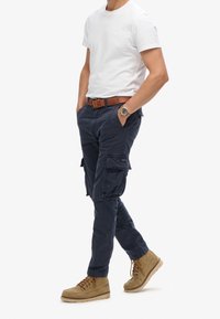 Marineblauwe cargobroek met zijzakken, gedragen met een wit T-shirt. Bruine riem en lichtbruine veterlaarzen maken de outfit compleet. Casual ontwerp.