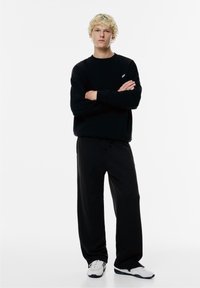 Zwarte sweatshirt met een ronde hals en logo, gecombineerd met zwarte sweatpants. Het model draagt witte sneakers met accenten. Effen achtergrond.