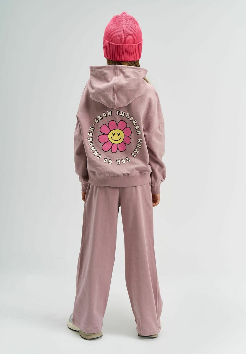 Un sweat à capuche rose avec un graphique de fleur et un texte au dos, associé à un pantalon ample assorti. Les deux sont faits d'un tissu doux. Un bonnet en tricot rose.